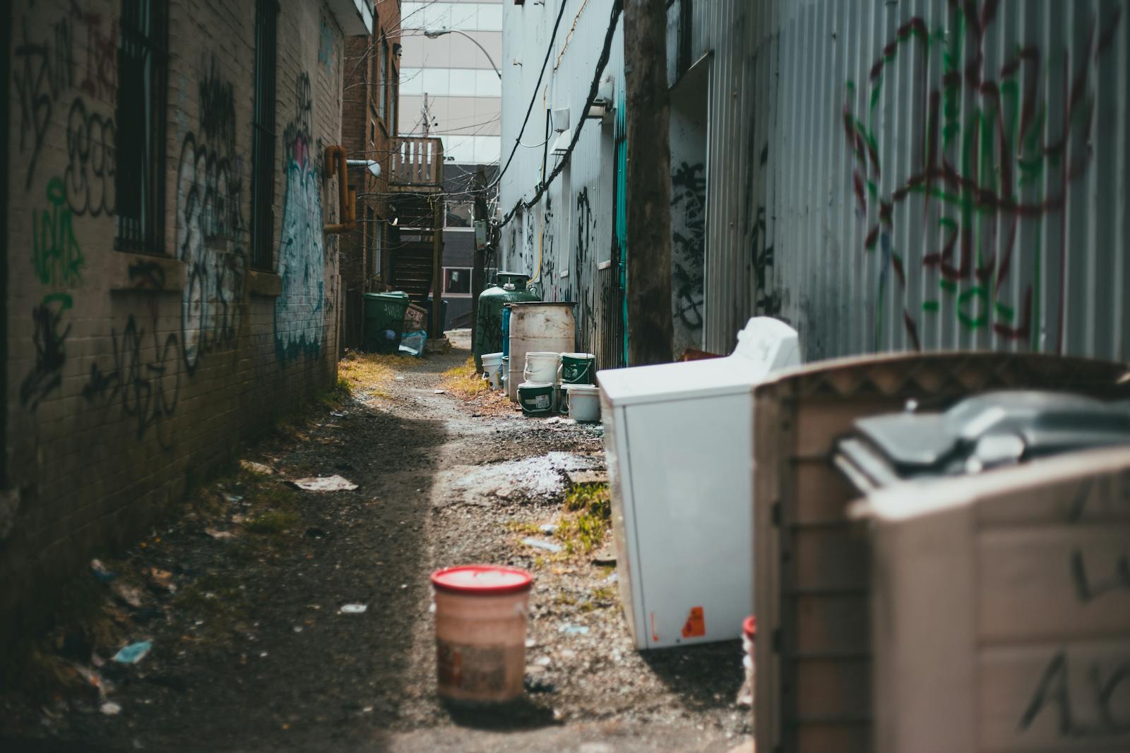 A narrow urban alley filled with scattered trash and graffiti-covered walls.