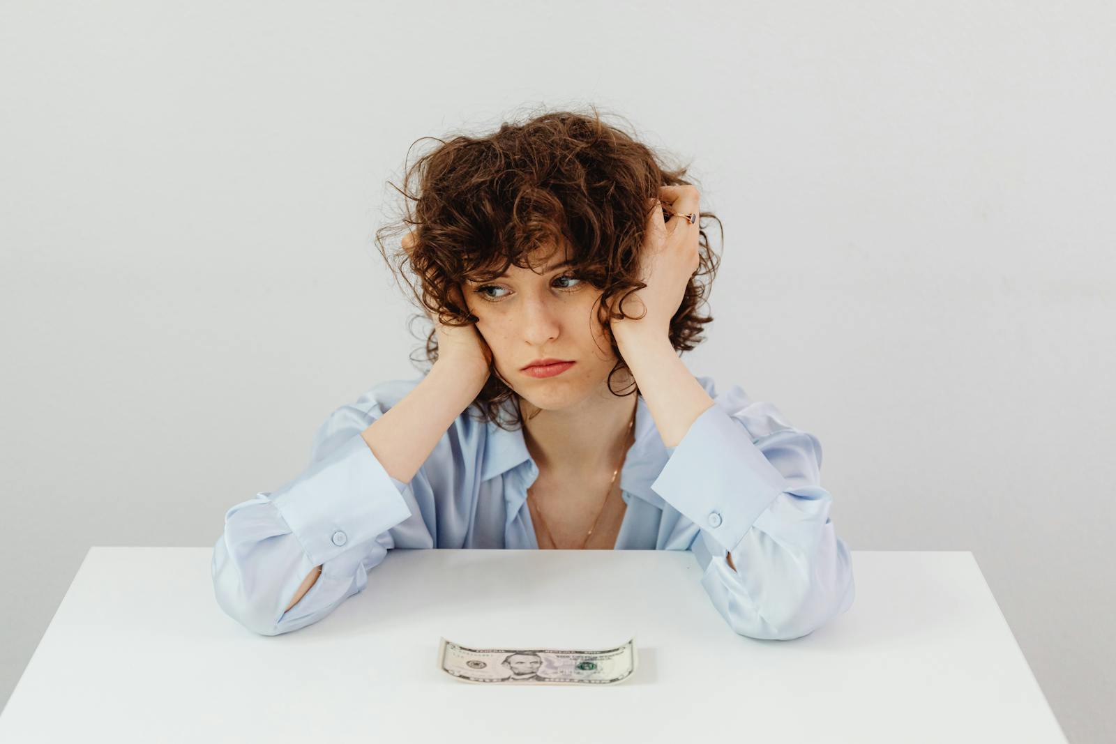 A woman in a blue blouse looks worried with a dollar bill on a table, symbolizing financial stress.
