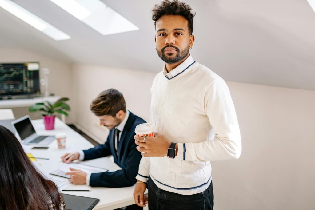 Confident businessman with coffee in a modern office setting with colleagues working nearby.