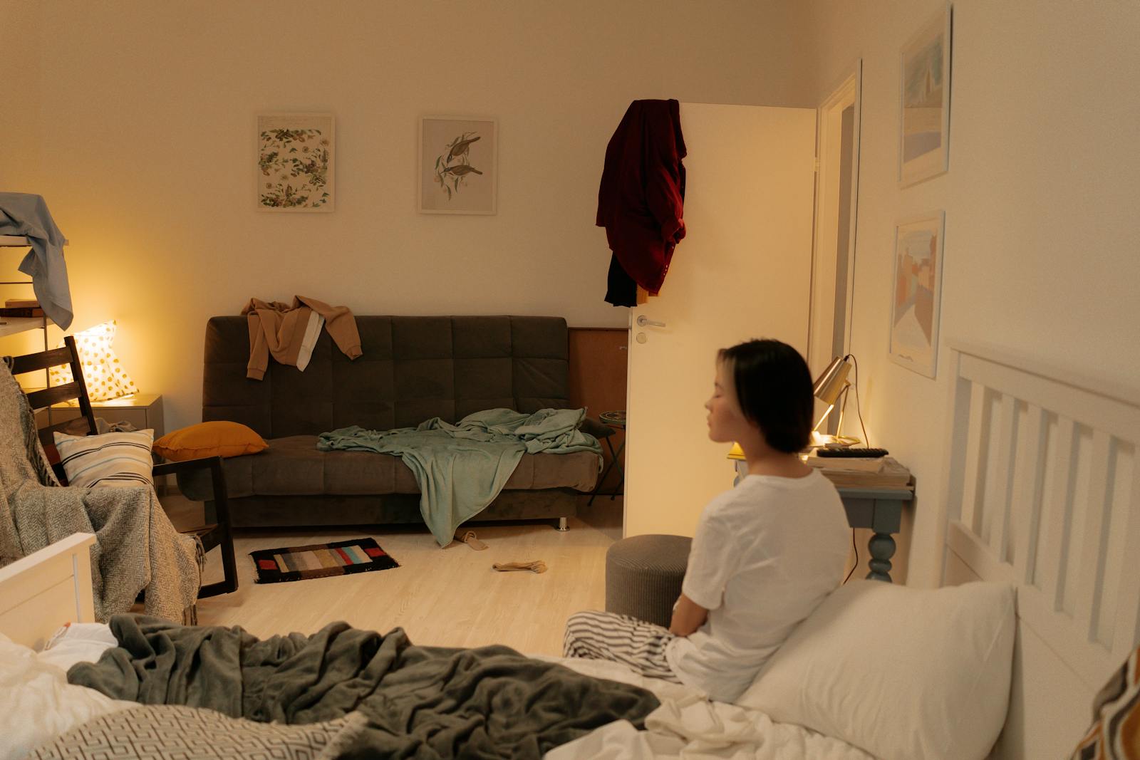 A woman sits on a bed in a messy bedroom with scattered clothes and furniture.