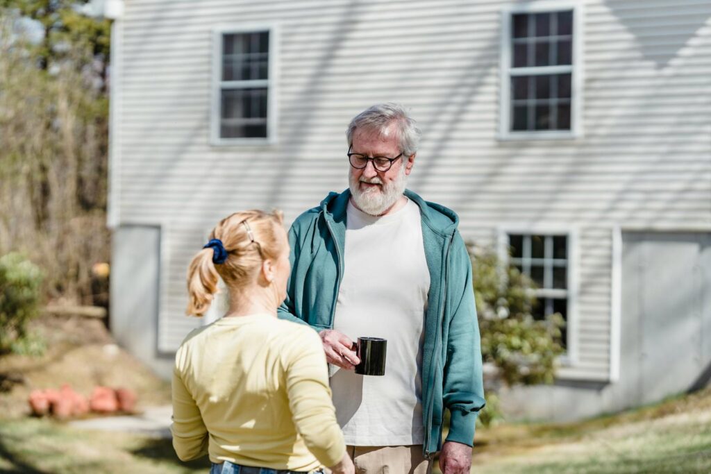 Elderly couple relaxing outdoors with mugs, having a peaceful conversation in the sunny yard.