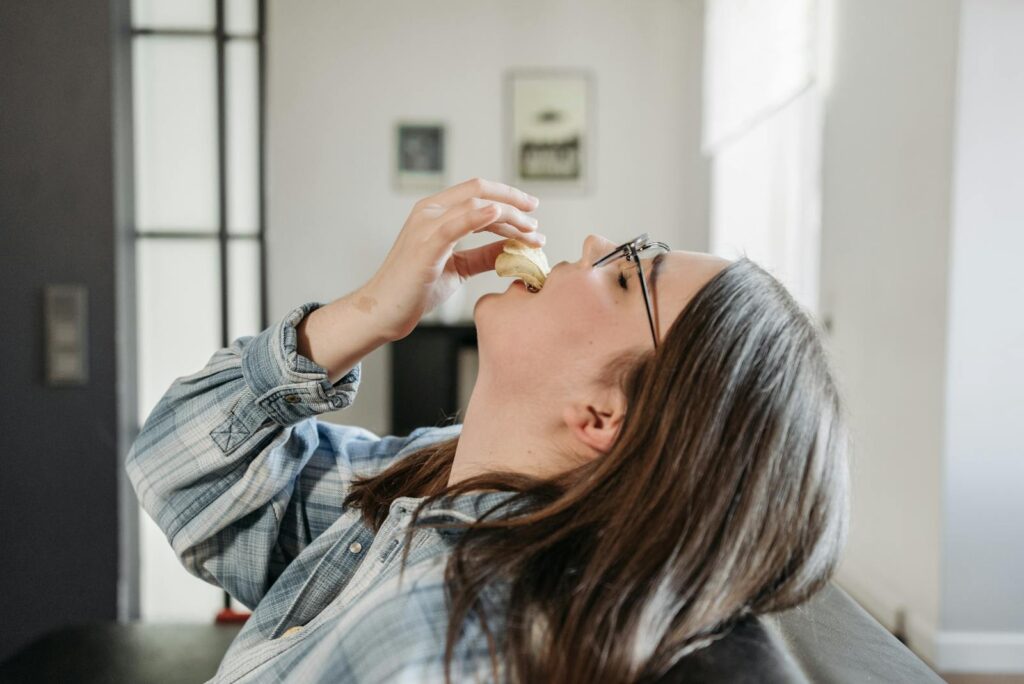Side view of a woman eating potato chips indoors, wearing plaid long sleeves and eyeglasses.