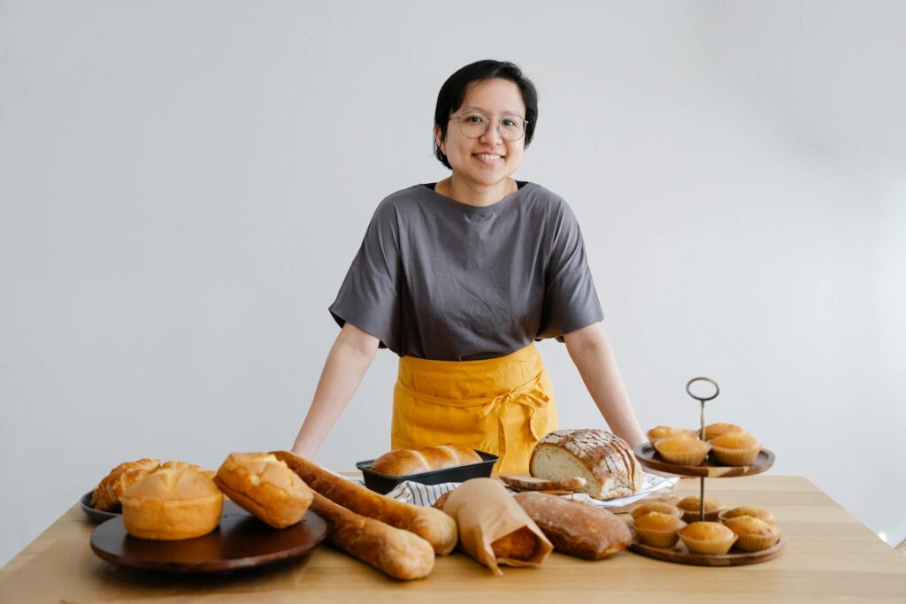Smiling Asian female baker showcasing a variety of fresh bread and pastries indoors.