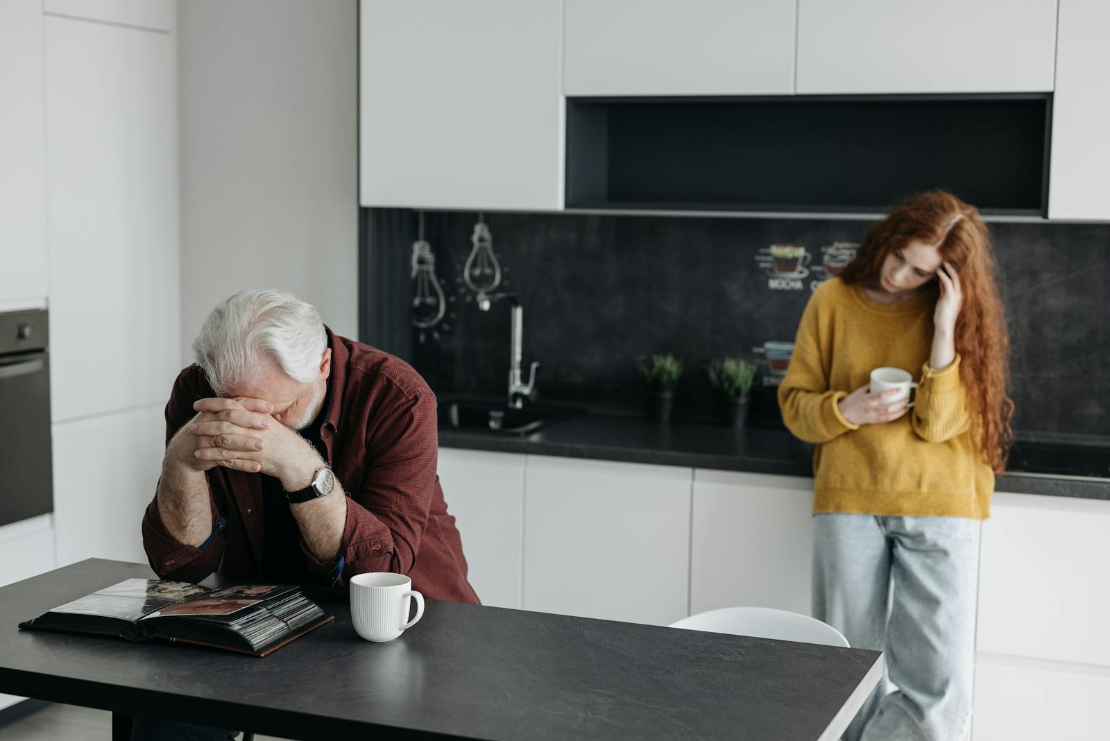 A man grieving at a kitchen table with a woman nearby, conveying emotions of loss and reflection.