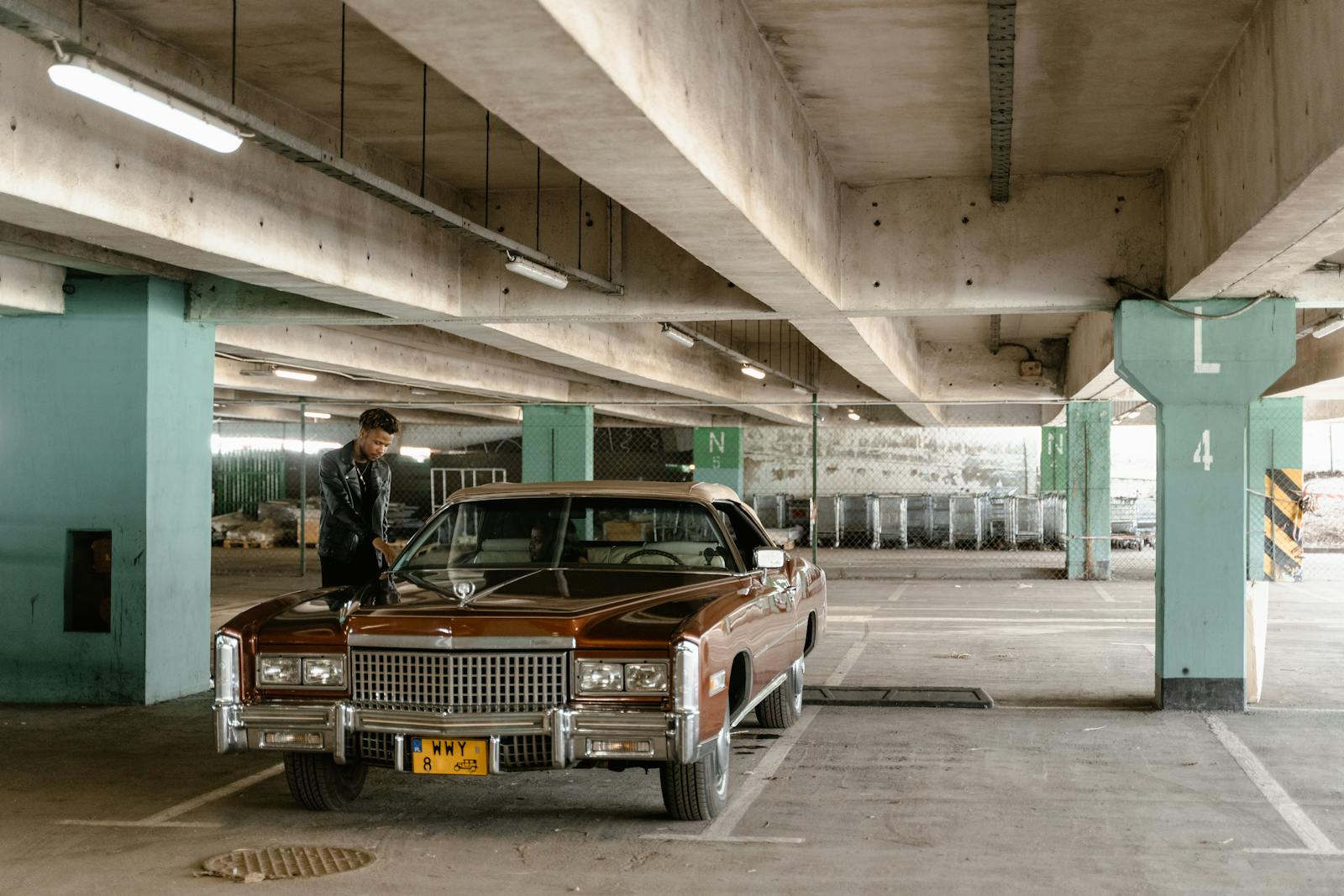 A man standing by a classic Cadillac in a multi-level parking garage.