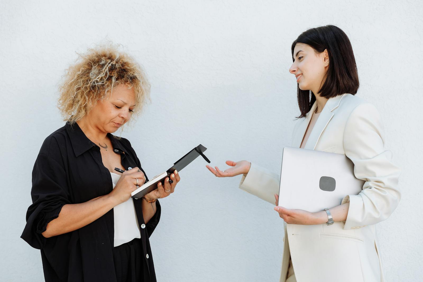 Two businesswomen engage in a discussion, one writing notes and the other presenting, symbolizing teamwork.