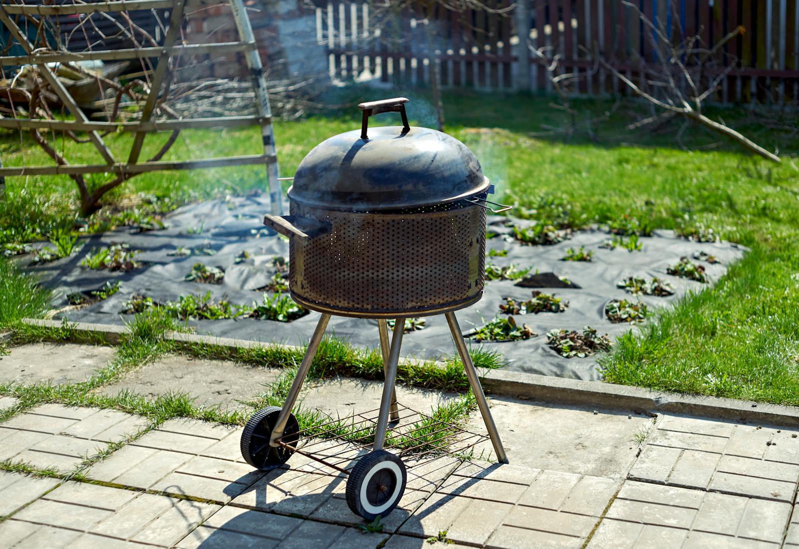 An outdoor charcoal grill with smoke in a sunny garden setting.
