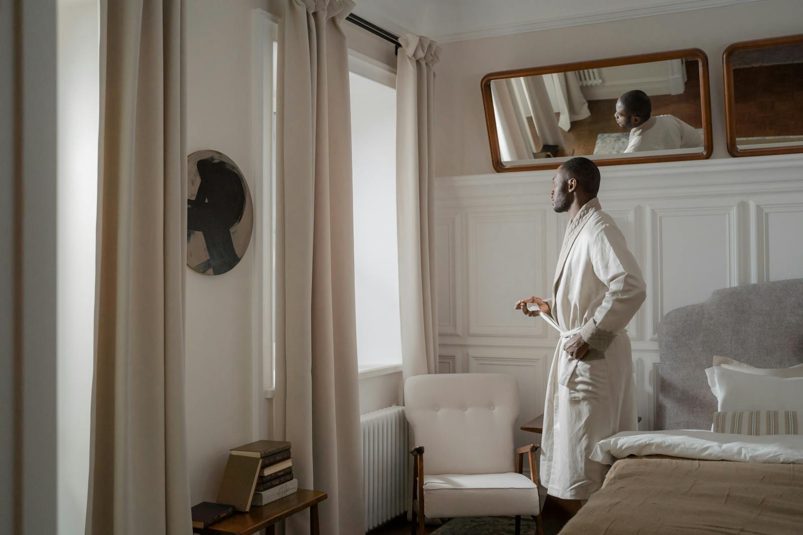A man in a bathrobe gazes out a window, reflecting in a serene bedroom.