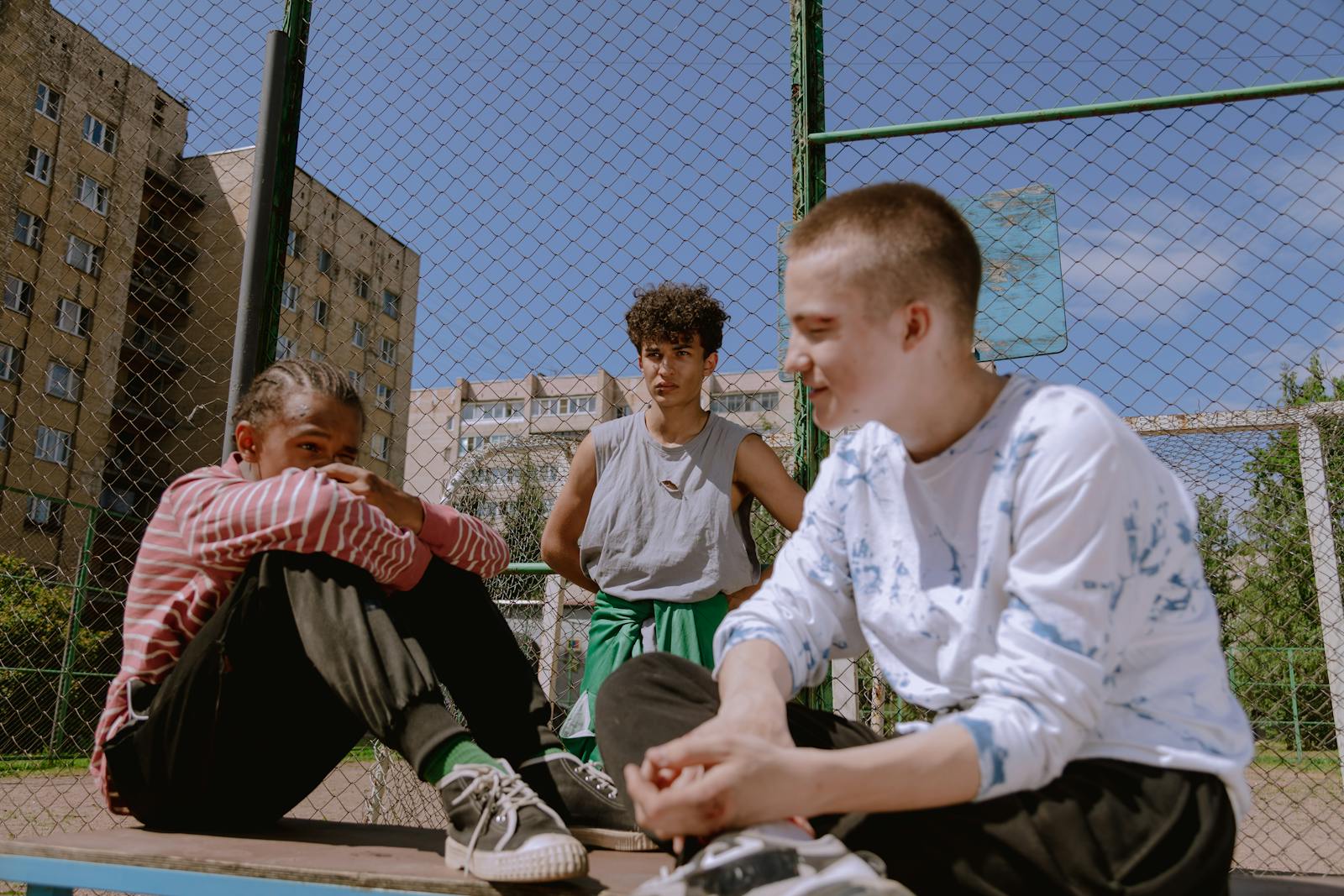 Three teenagers sitting and relaxing on an outdoor basketball court on a sunny day.