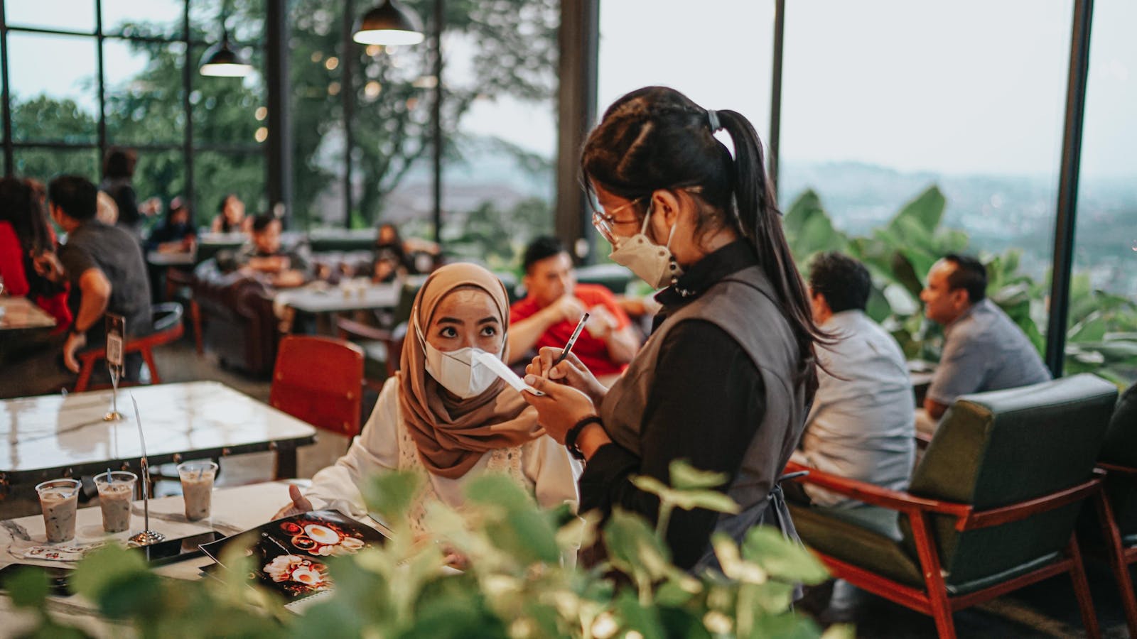 A waitress wearing a face mask takes an order from a customer in a bustling café, highlighting the new normal in dining.