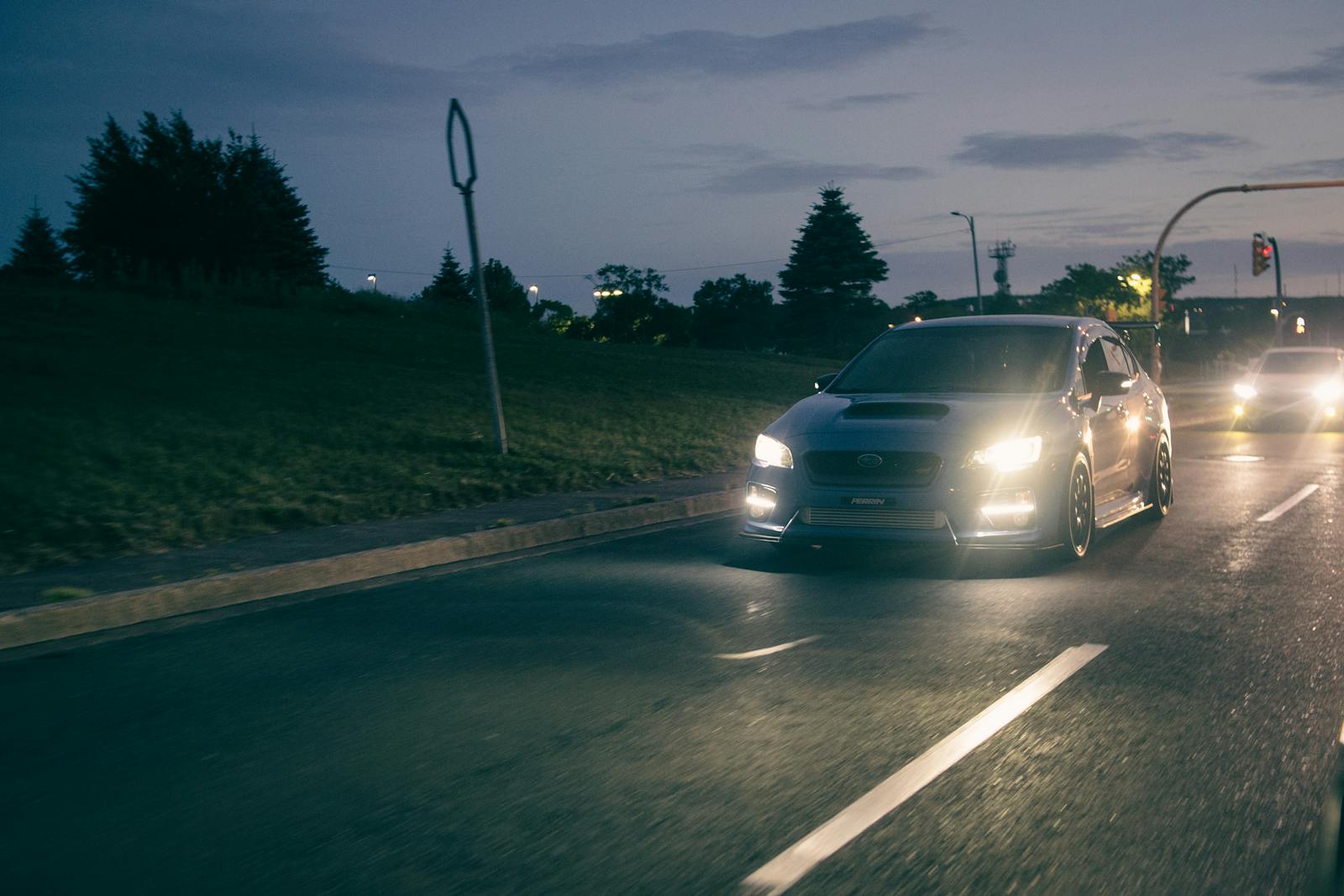A sleek Subaru WRX drives on a city street at dusk, headlights on, capturing the essence of night travel.