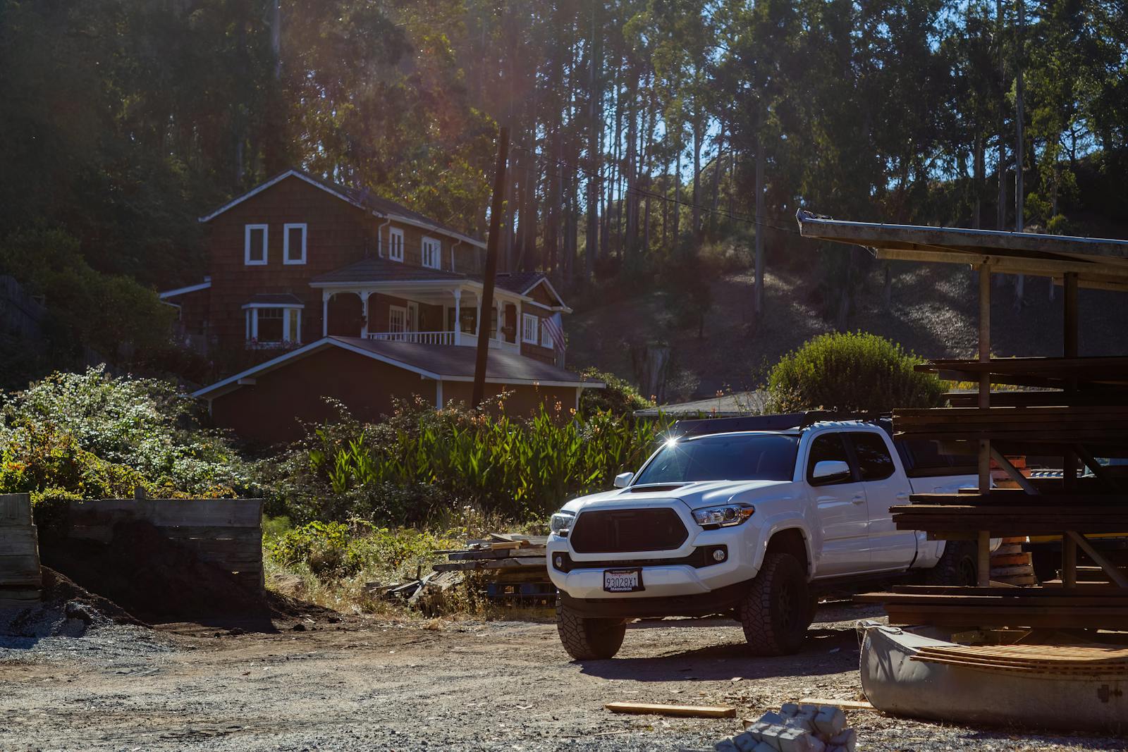 White pickup truck parked on a rustic countryside driveway near a wooden house under sunlight.