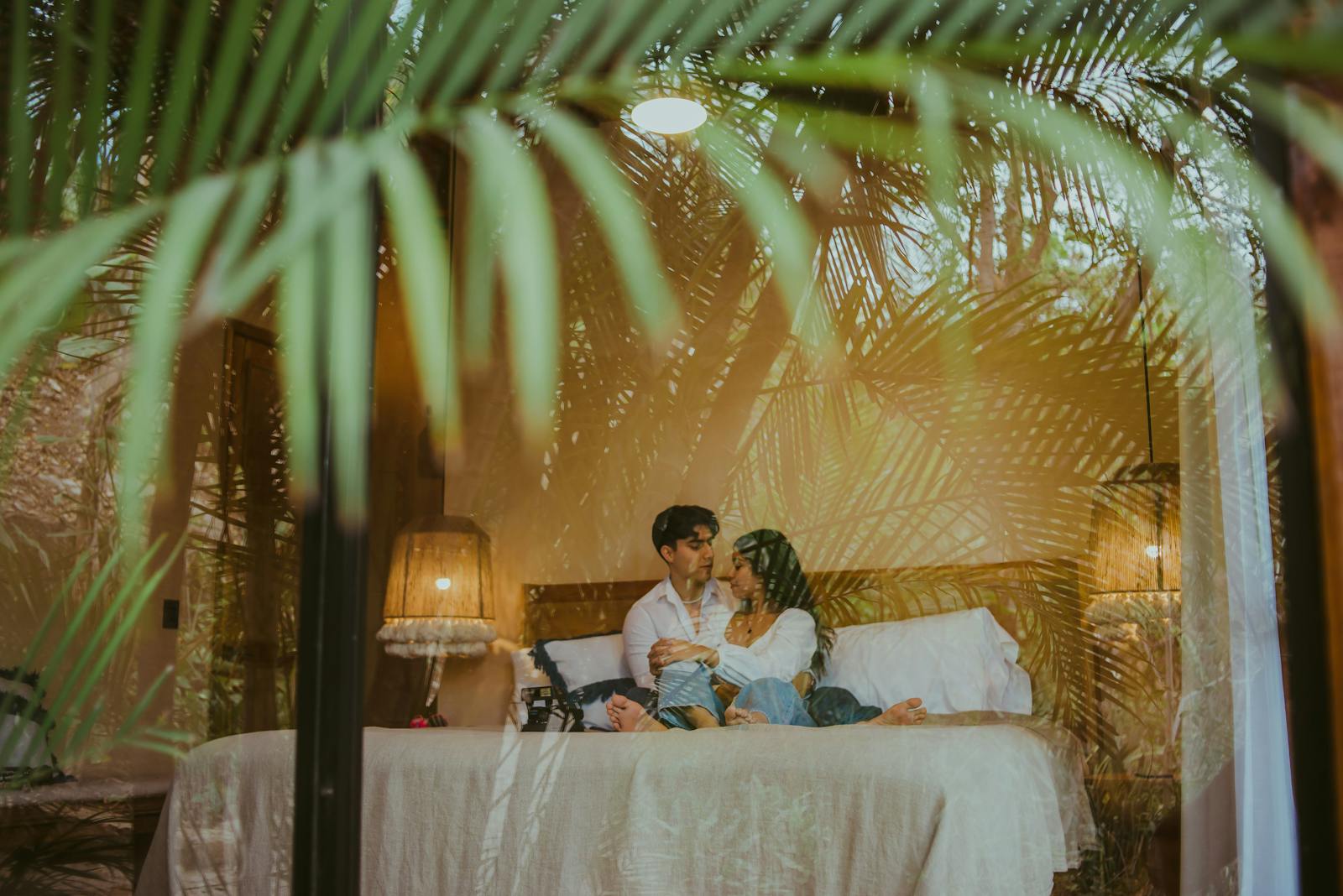 A couple lovingly embraces on a bed in a cozy, illuminated hotel room.
