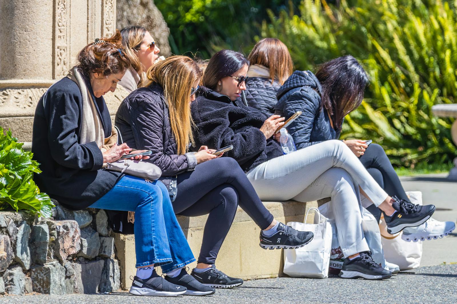 Five women sitting outdoors, using smartphones, enjoying sunny day in park.