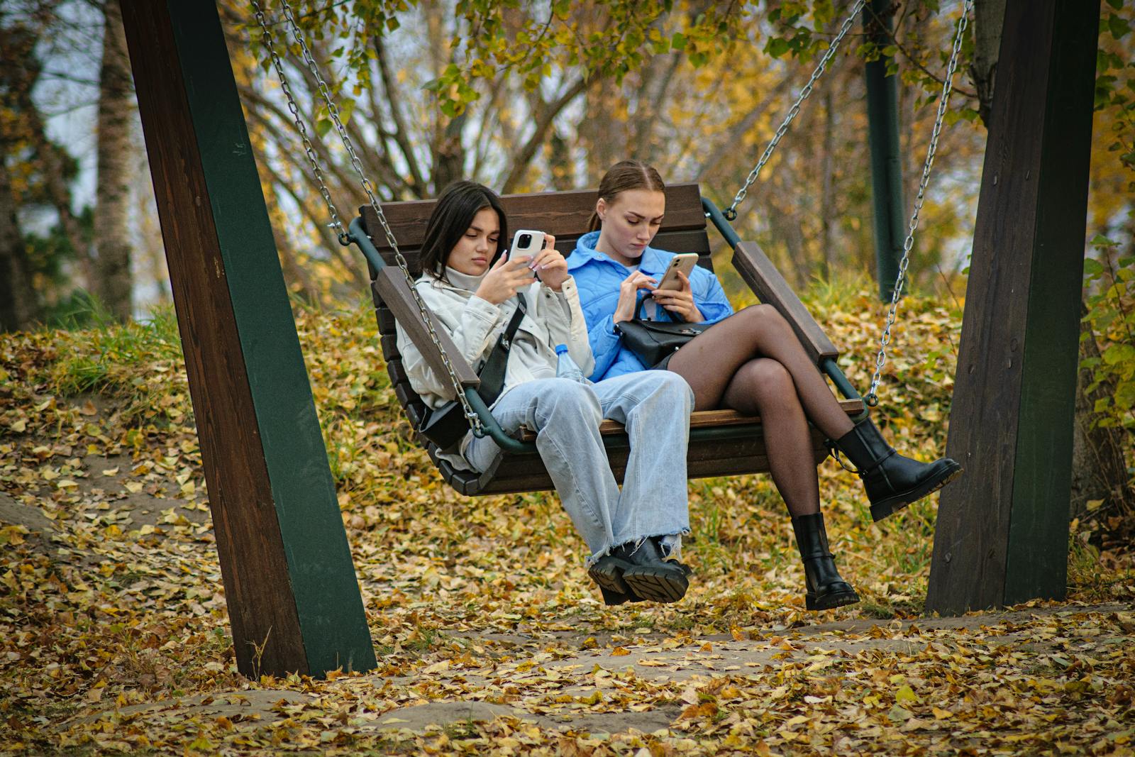 Two young women sitting on a swing using smartphones, surrounded by autumn leaves in a park.