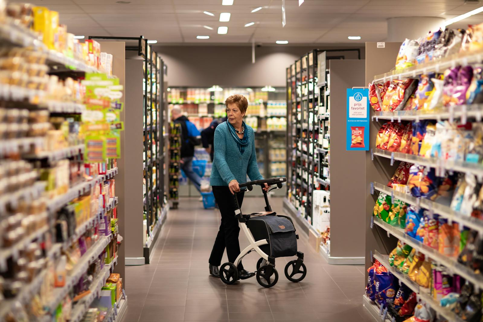 Elderly woman using a rollator for support while shopping in a supermarket aisle.