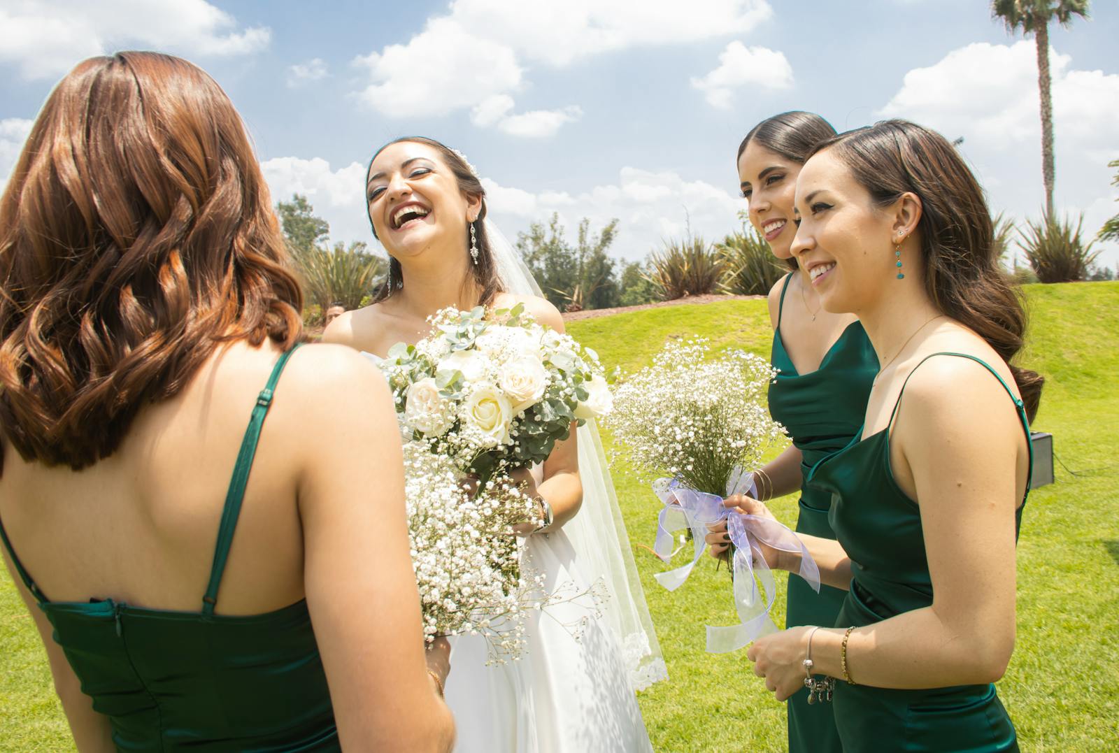 A happy bride shares a laugh with her bridesmaids outdoors in a lush green setting in León, Mexico.