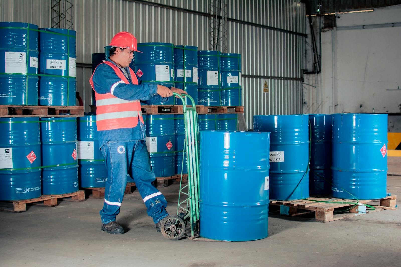 Industrial worker with a hand truck moving blue barrels in a warehouse setting.