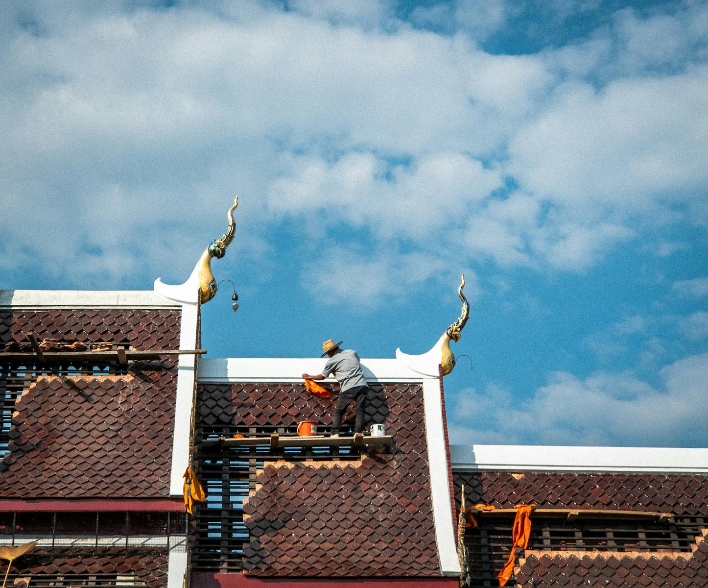 A craftsman repairing a temple roof against a vibrant sky in Chiang Mai, Thailand.