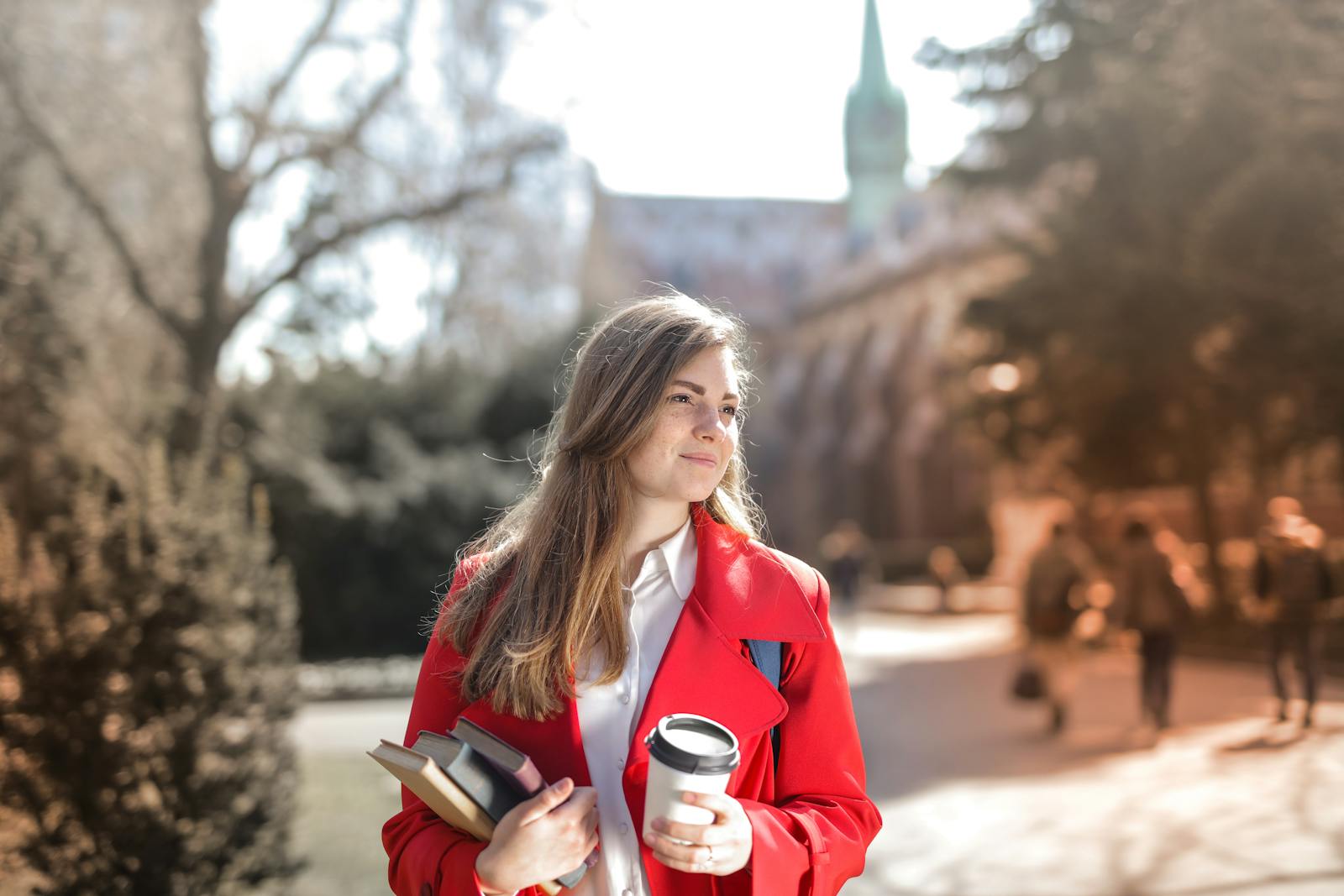Smiling woman in red coat with coffee and books, enjoying leisure time outdoors on a university campus.