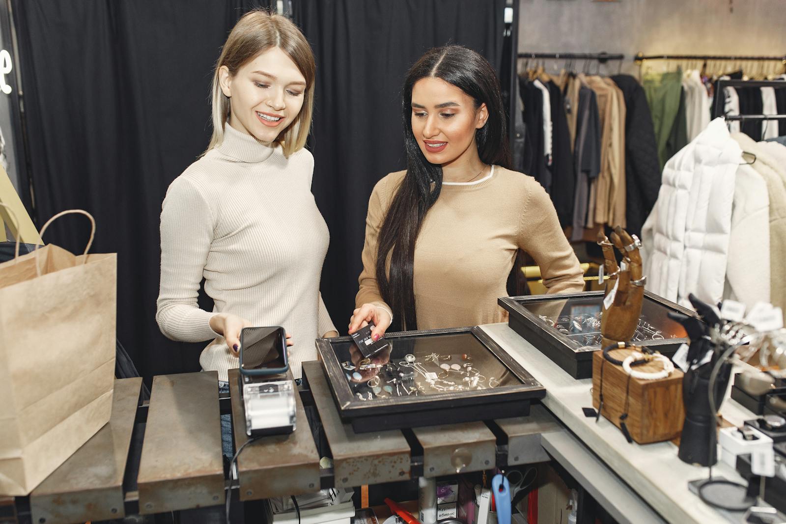 Young cheerful women using smartphone and credit card while paying for purchases and toothy smiling while standing at counter table near showcase with accessories