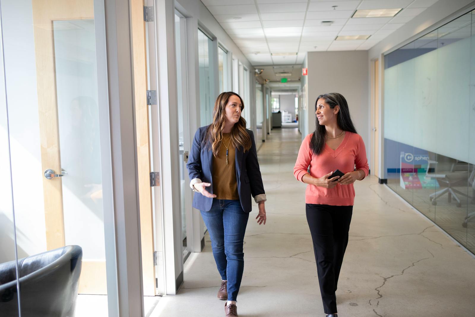 Two professional women engaged in conversation while walking through a modern office corridor.