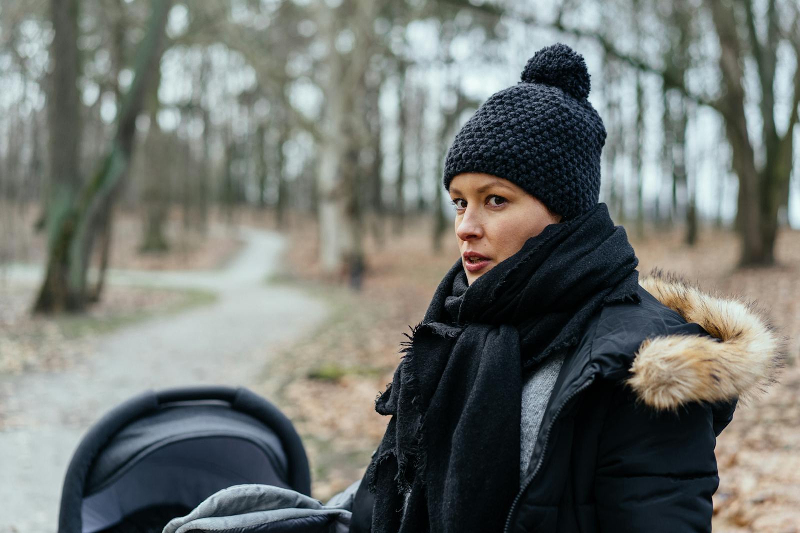 A woman in a beanie and scarf walking with a stroller through a park on a winter day.