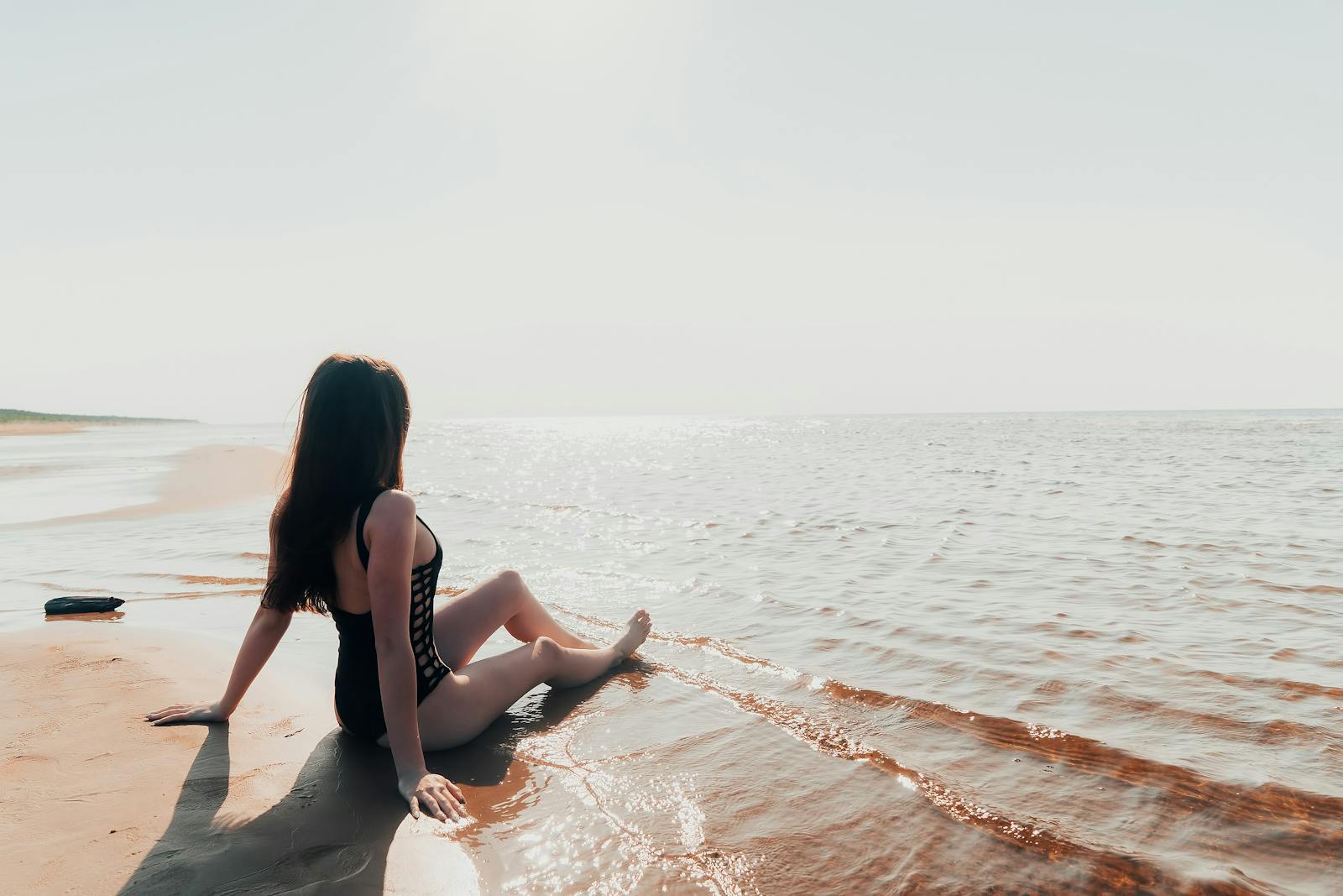 A woman in a swimsuit sits peacefully on a sandy beach by the ocean, enjoying a sunny day.