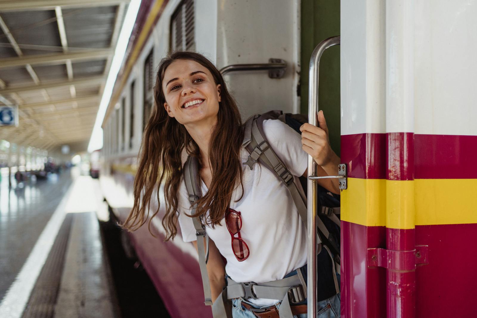 A cheerful woman with a backpack smiling while leaning out of a train at a station.