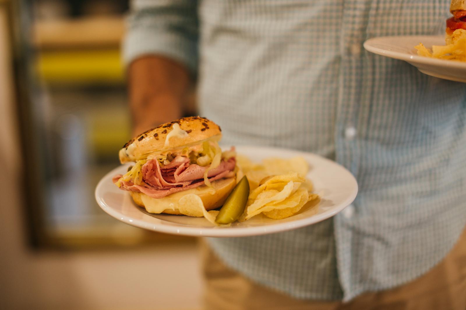 Plate with a tasty sandwich, chips, and a pickle held by a man indoors.