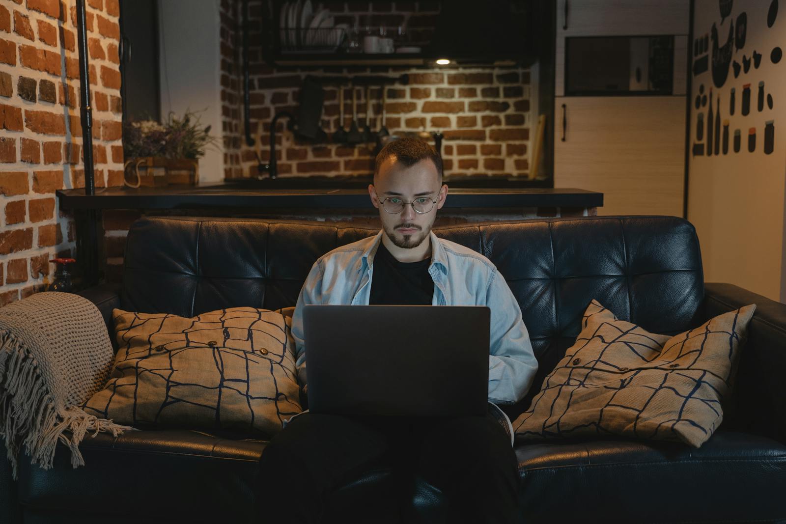 Man working remotely on a laptop in a cozy, stylish apartment with exposed brick walls and modern decor.