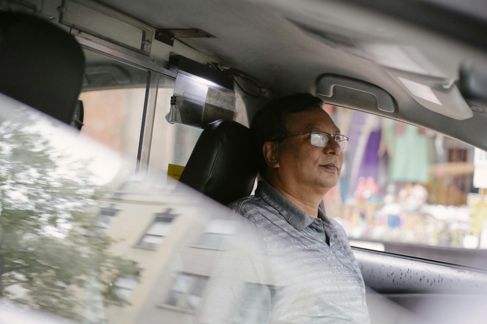 Side view of Asian middle aged male in eyeglasses sitting on driver seat in automobile with reflection of building on half opened window