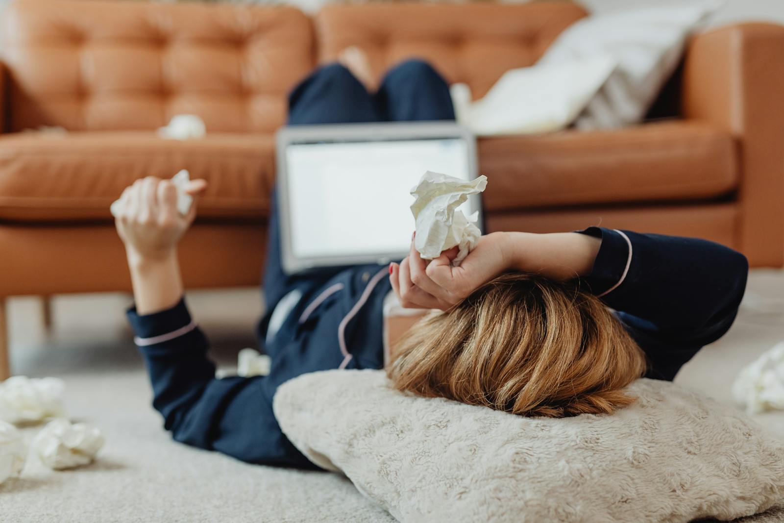A woman in pajamas lies on the floor next to a laptop, surrounded by tissues, depicting a sick day at home.