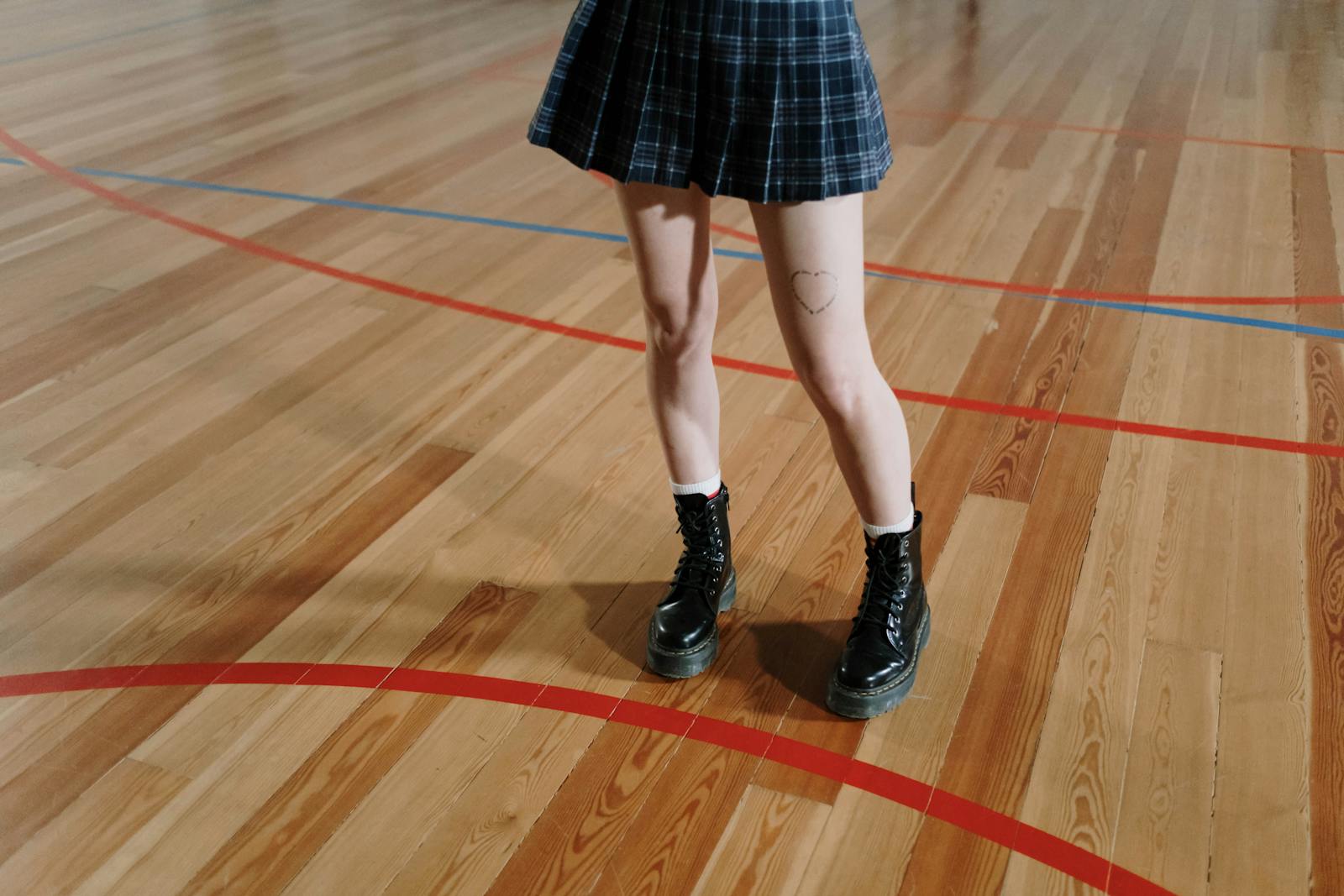 Teenage girl stands on gym floor wearing boots and checkered skirt, showcasing a stylish look.