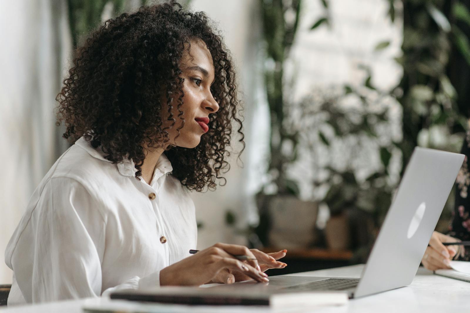 Young woman with curly hair typing on laptop in a bright, plant-filled office space.