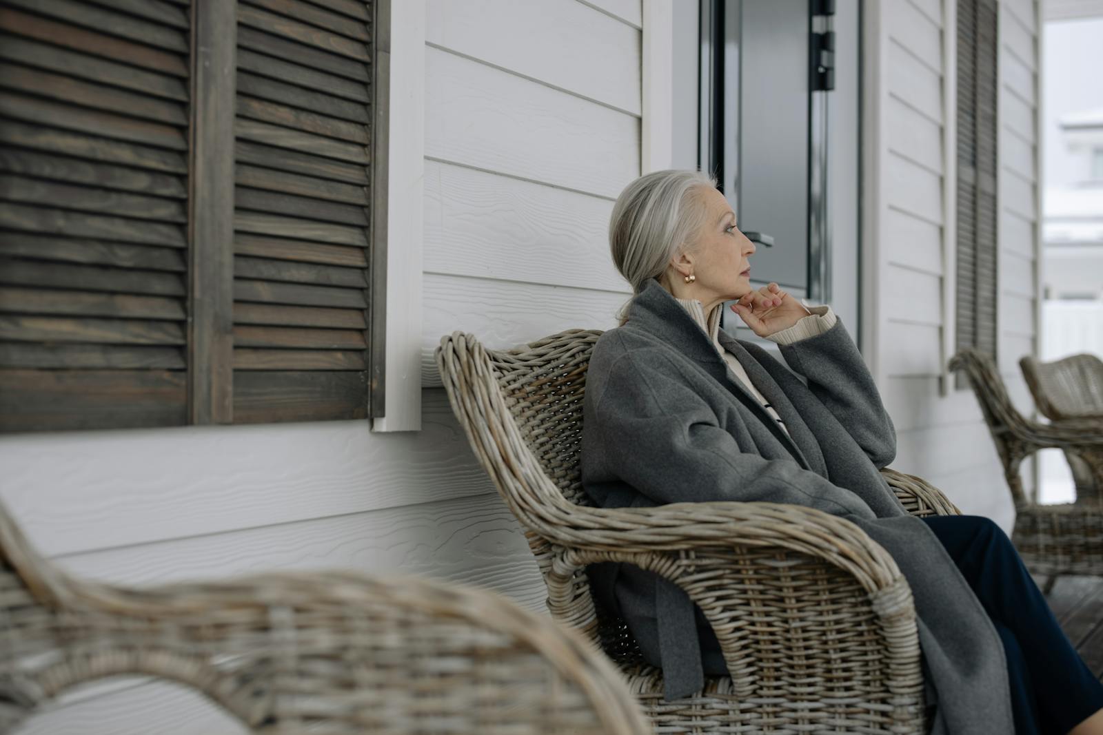 Elderly woman sitting in a cozy wicker chair on a porch, reflecting in winter wear.