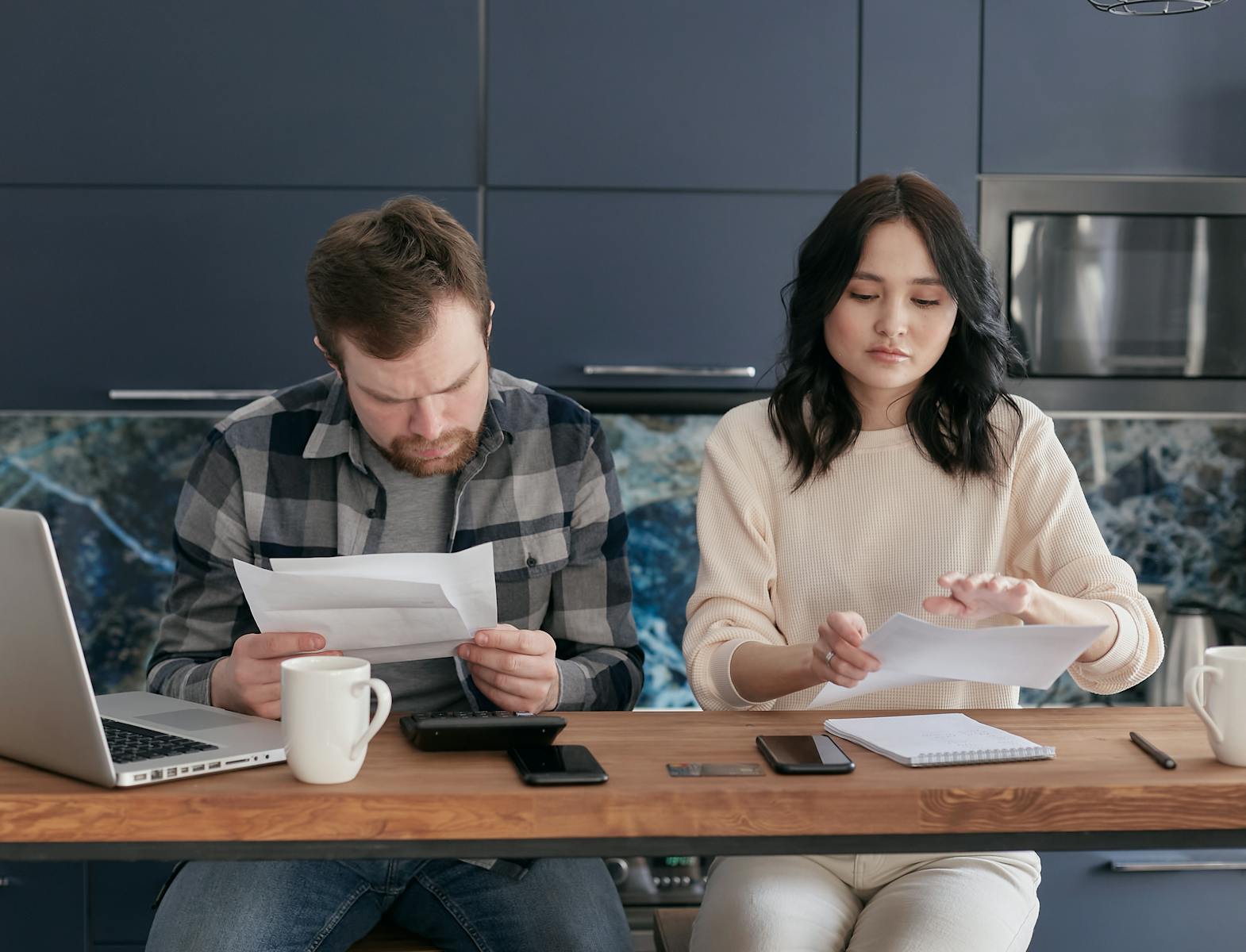 A couple reviews bills and manages finances on a kitchen table, looking concerned.