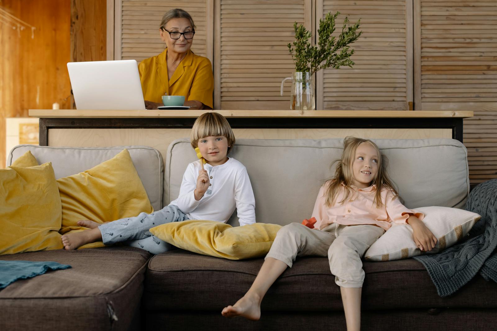 Elderly woman working on a laptop at home while grandchildren relax on the couch.