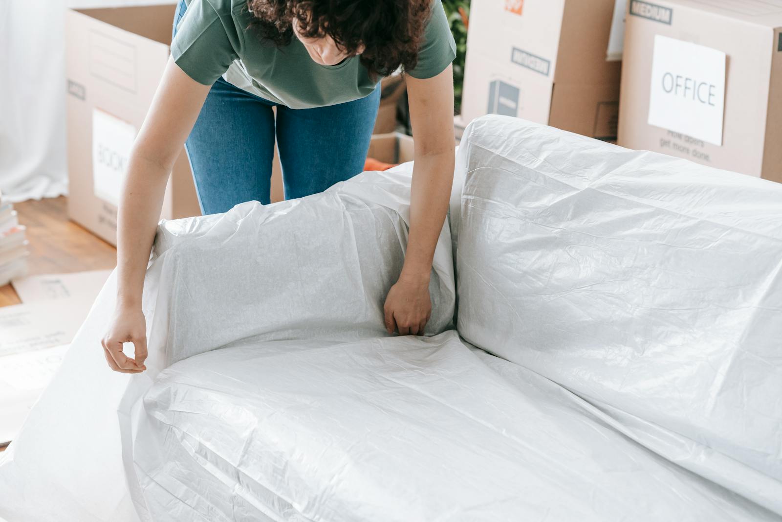 A woman carefully wraps a sofa in protective material while packing boxes in a bright room.