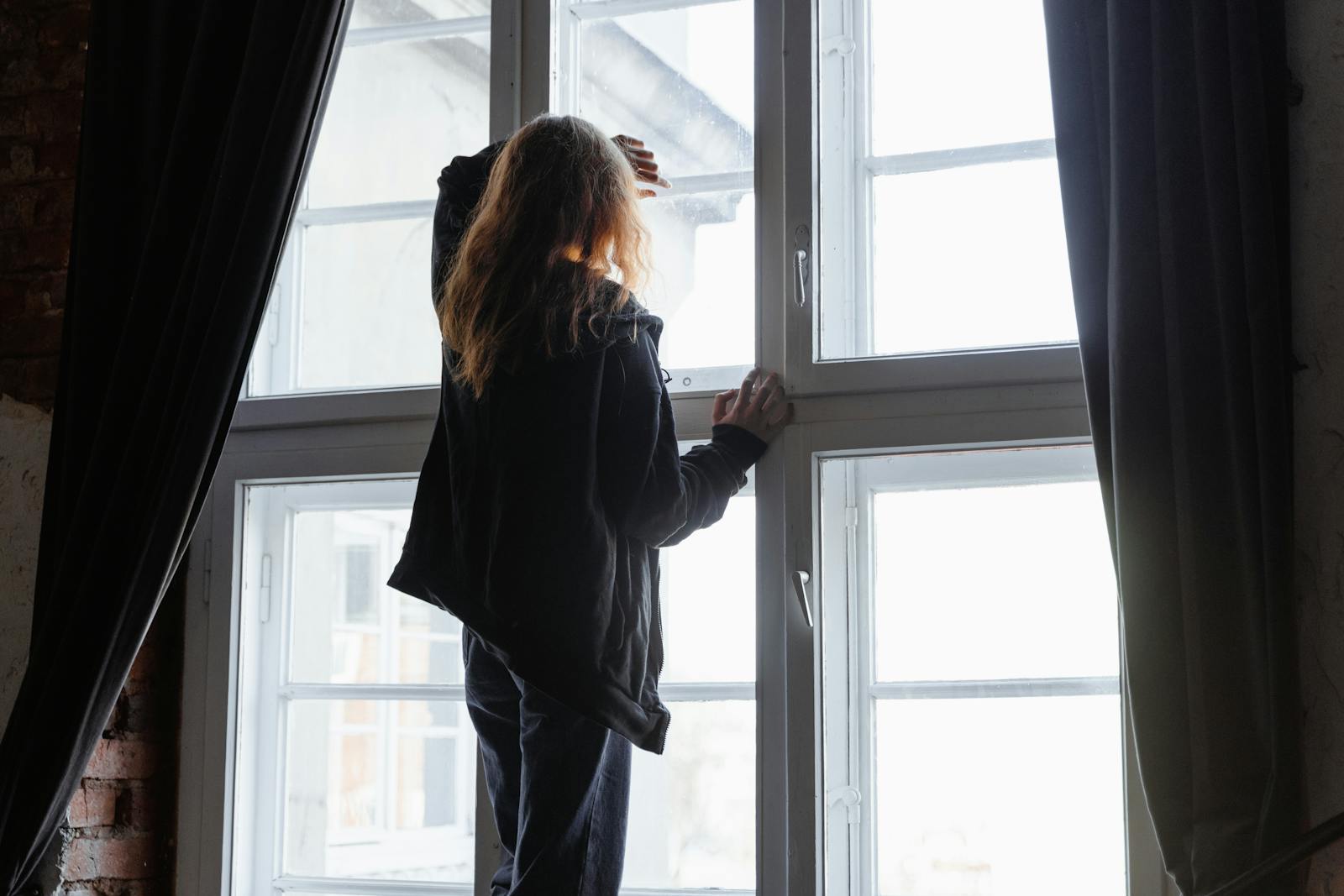A woman stands by a window, lost in thought, with natural light streaming in.