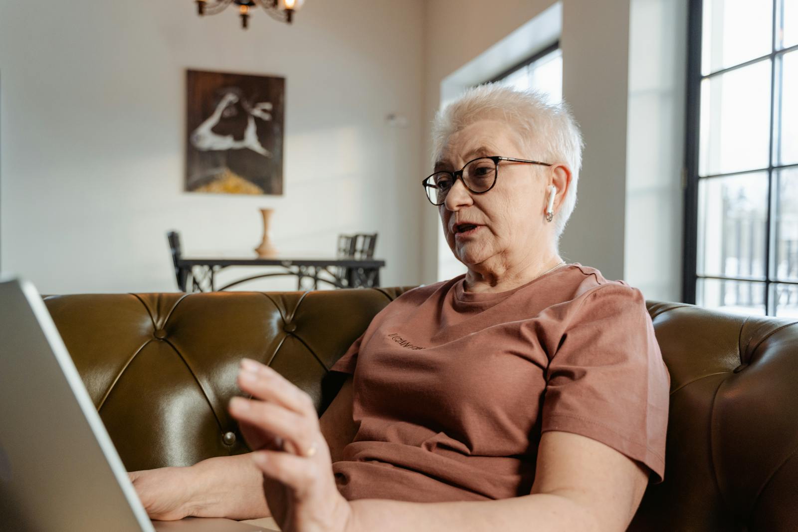 Elderly woman engaging with technology while sitting on a sofa, using a laptop and wearing earbuds.