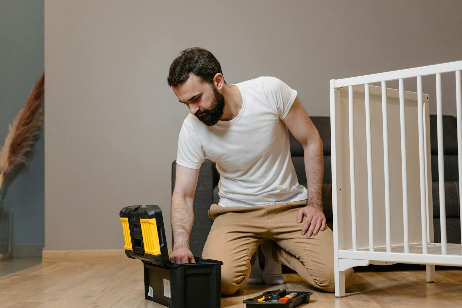 A man kneels indoors assembling a baby crib using a toolbox, wearing a white shirt and beige pants.