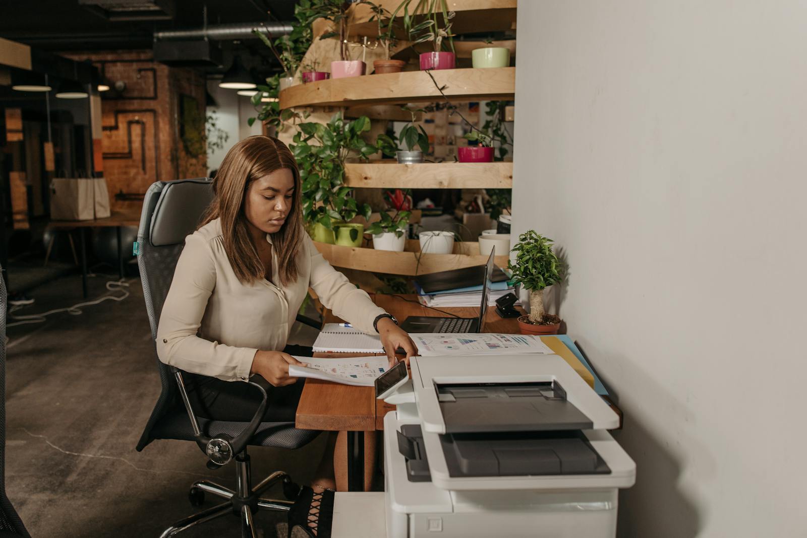 A focused woman works at a desk in a modern office environment, surrounded by plants.