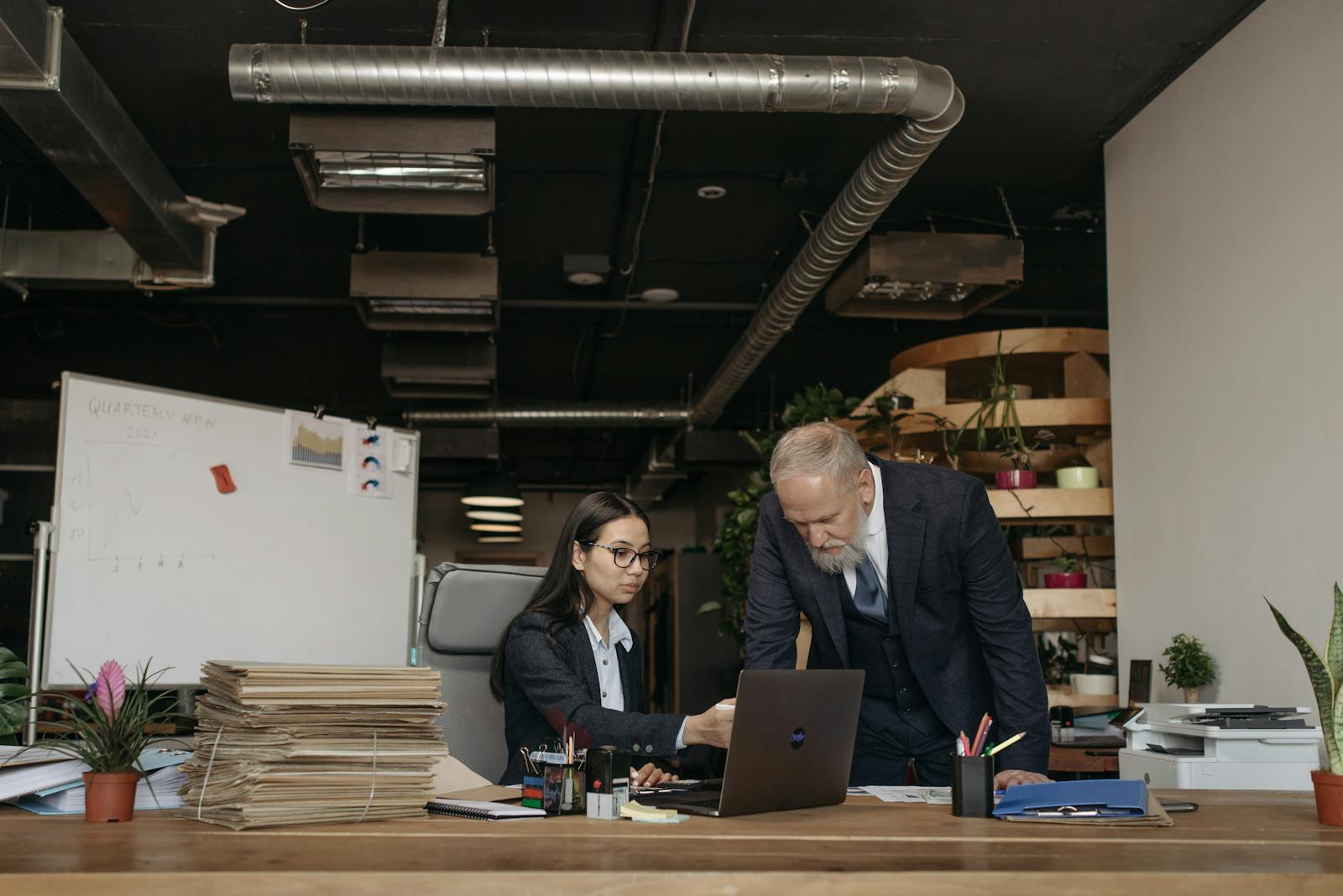 A senior man and woman collaborate on a laptop in a modern office setting.