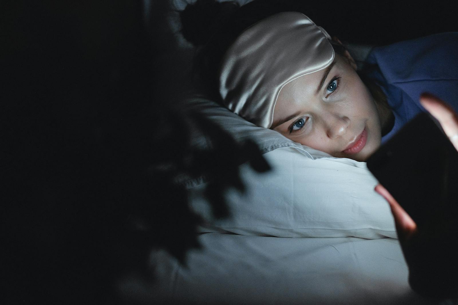 A woman in bed wearing a sleep mask uses her phone at night, creating a cozy ambiance.