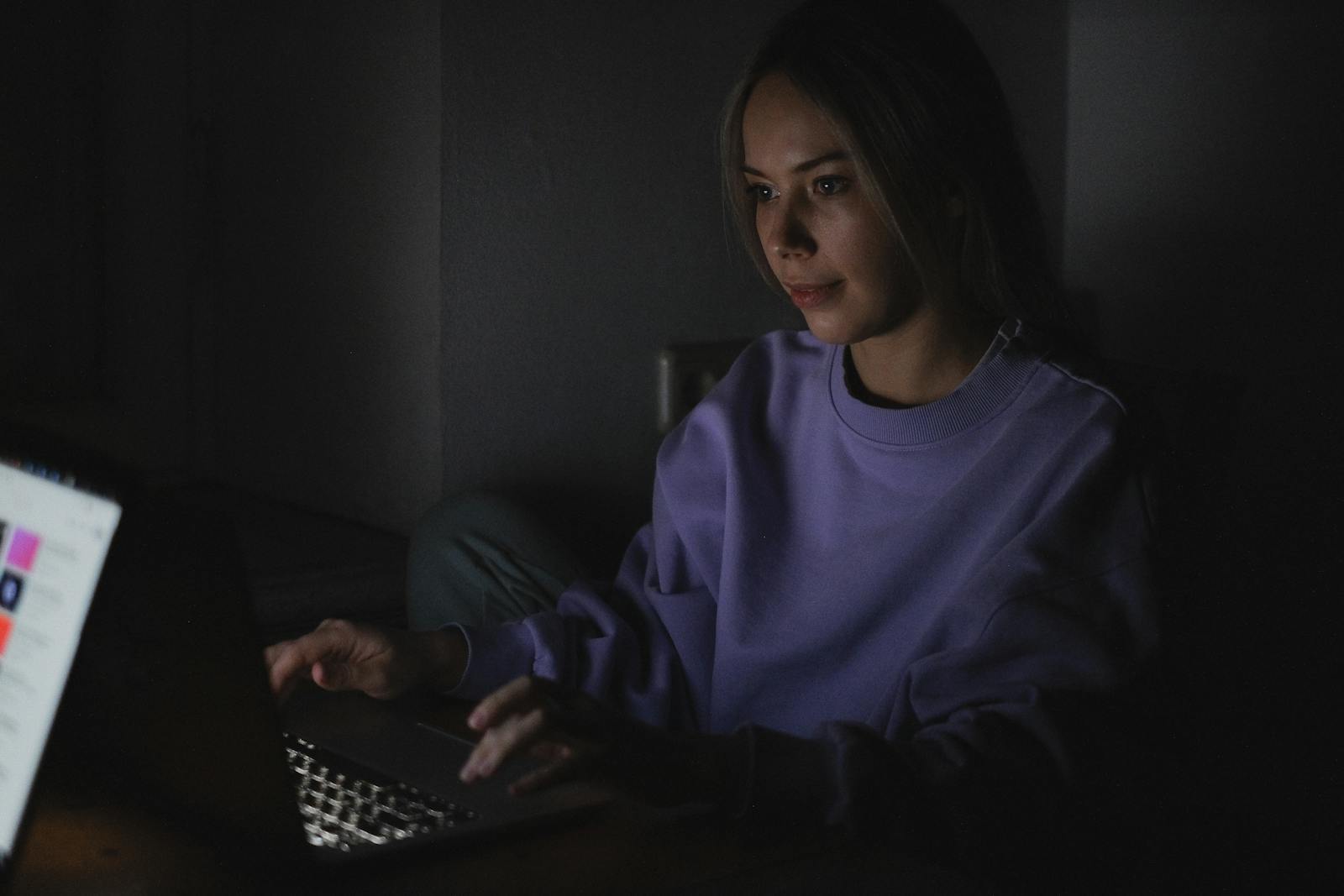 A woman wears a sweater and uses a laptop in a dark room, creating a focused and calm atmosphere.