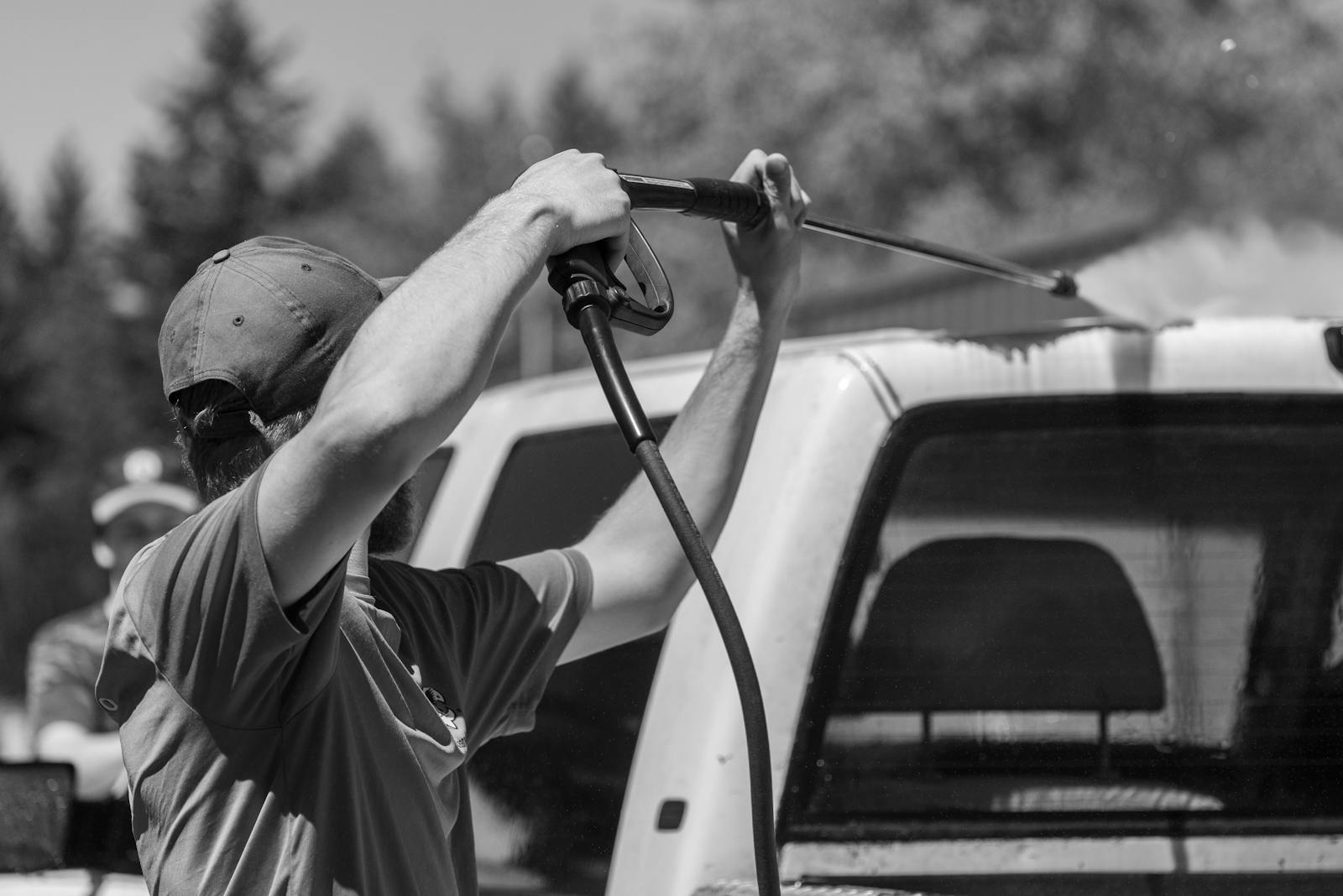 Black and white image of a man using a power spray on a truck outdoors.