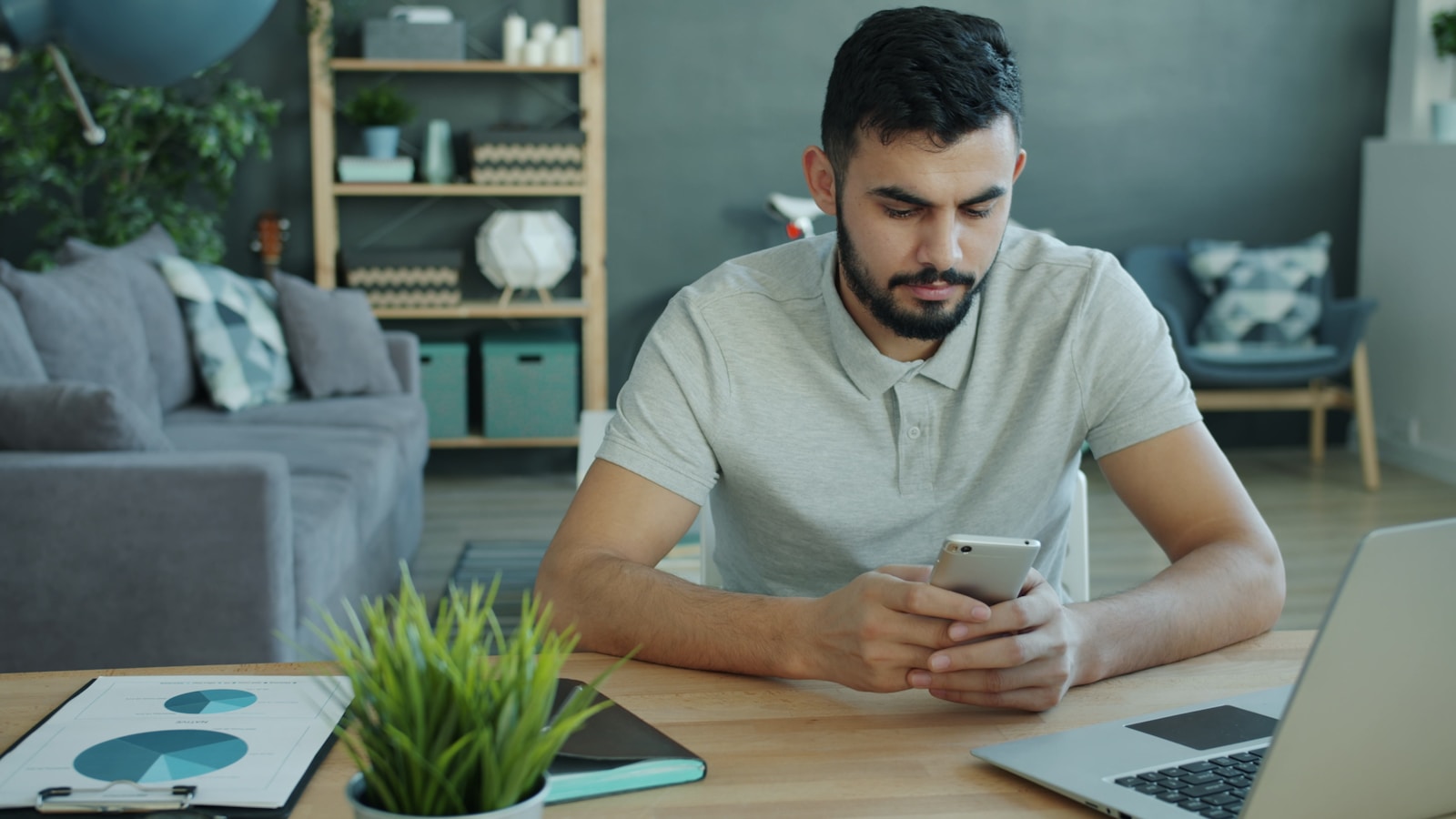 Man using smartphone at desk with laptop and charts.