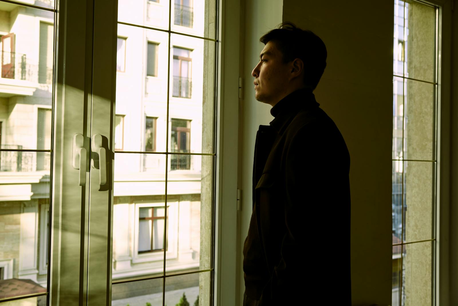 Contemplative man standing by a window indoors, gazing outside.