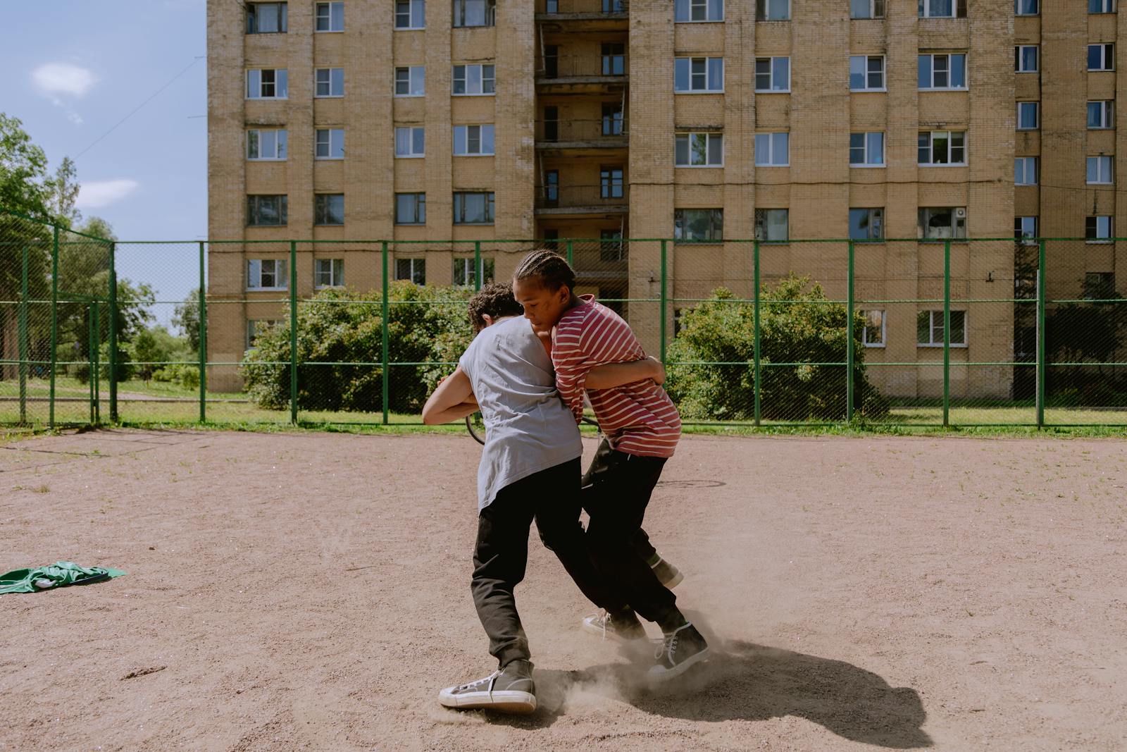 Two teenage boys engaged in playful wrestling on a dirt playground, outdoor sunny day.